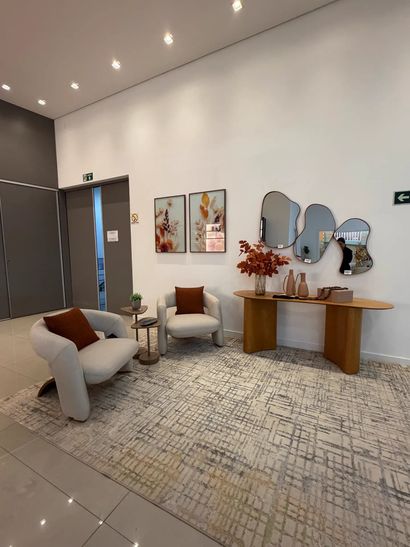 Hall do showroom com poltronas claras, aparador em madeira curva e espelhos orgânicos