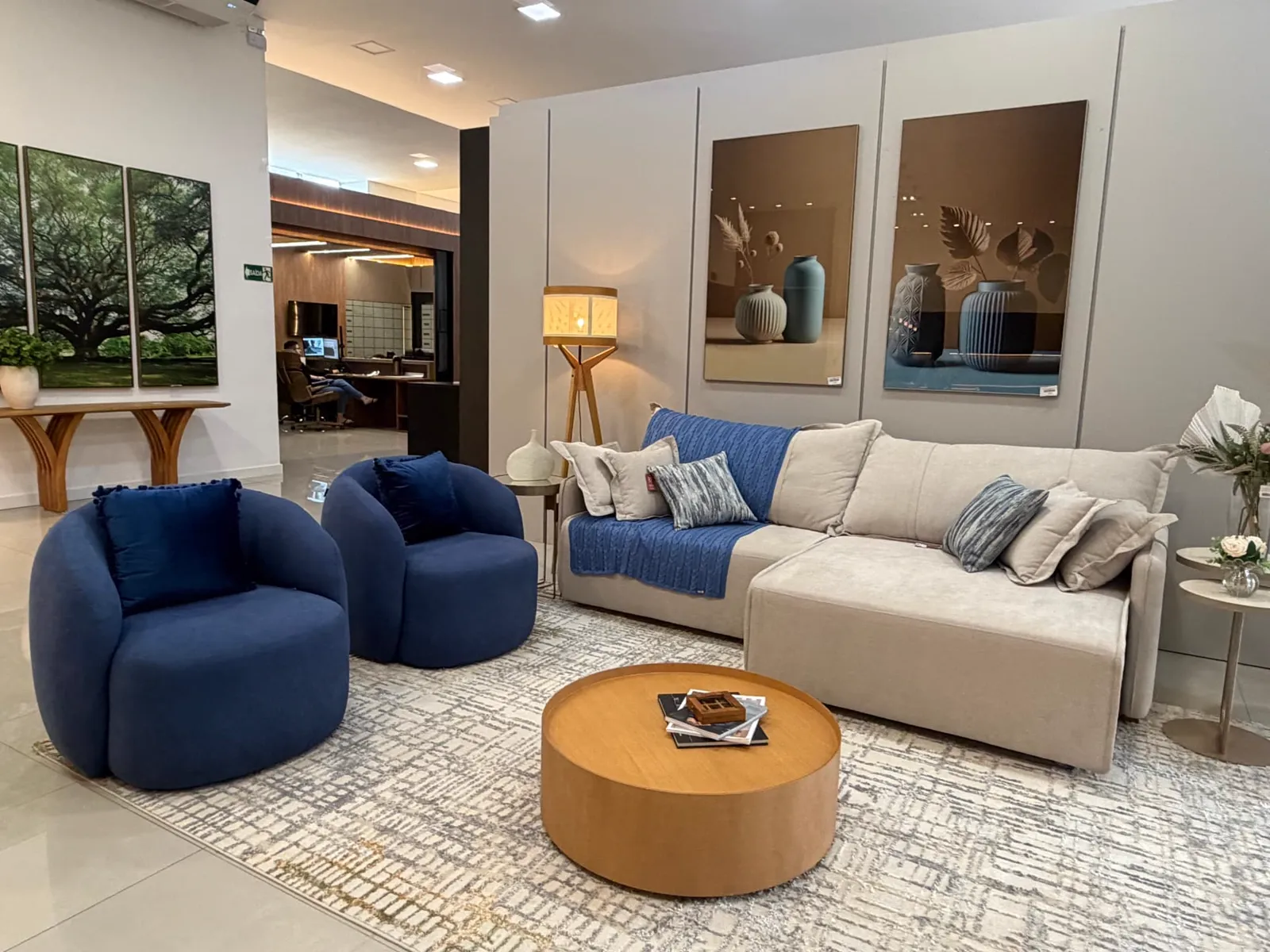 Estar do showroom com sofá claro, poltronas azuis e mesa de centro redonda em madeira